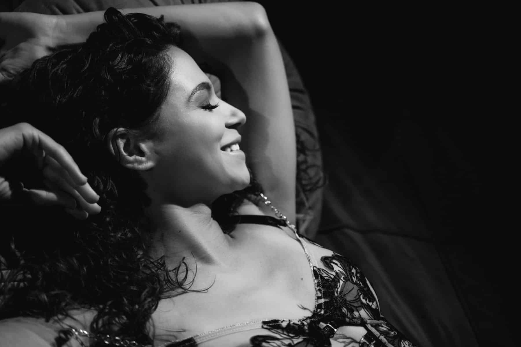 Black and white photo during a tattooed boudoir session of a woman laying on a bed, closeup, smiling with eyes closed because of her empowering and uplifting experience with Allebach Photography