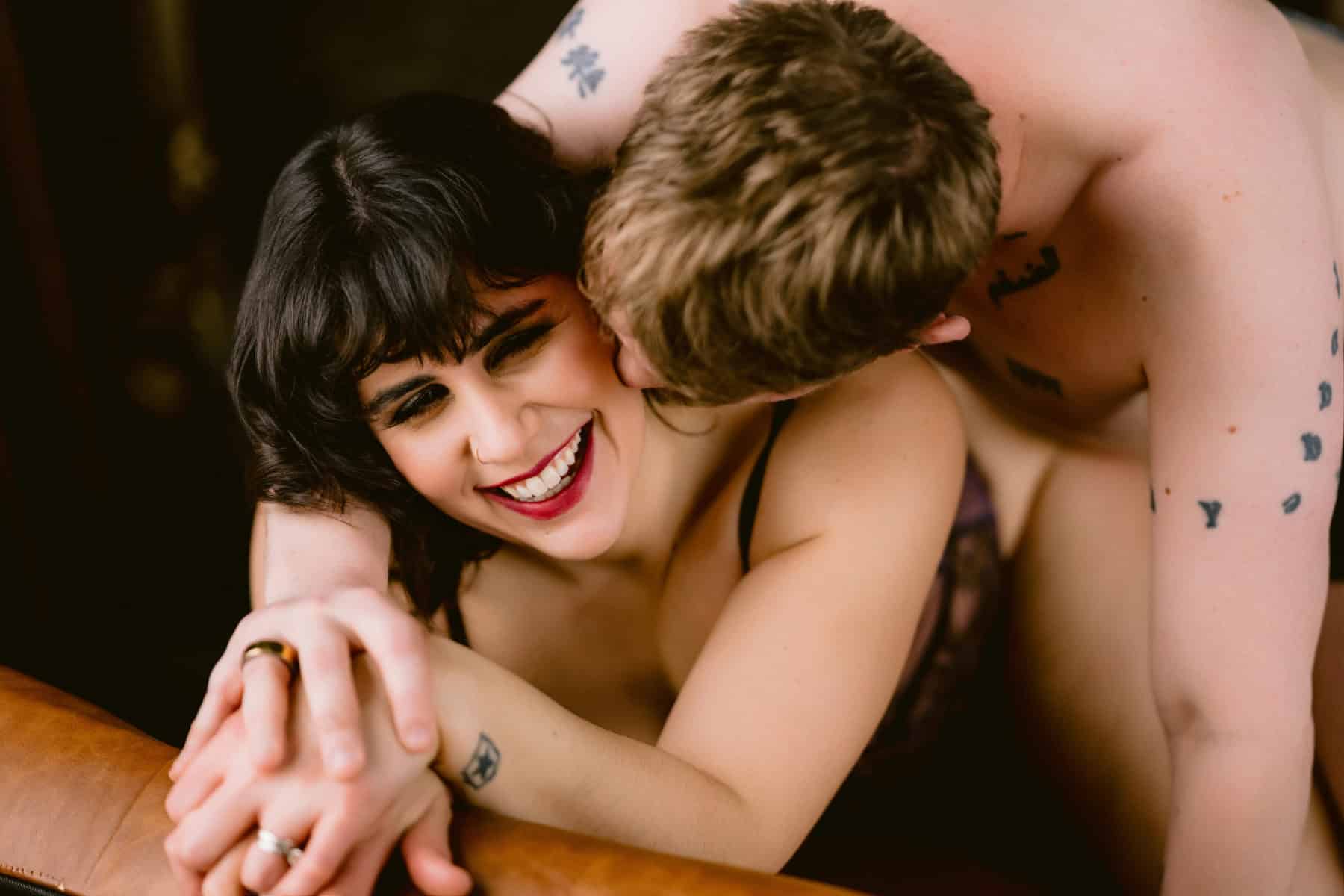 Closeup photo of a straight couple, woman kneeling with her husband behind her kissing her cheek, while she smiles with bright red lipstick, during a couples boudoir anniversary session with allebach photgraphy