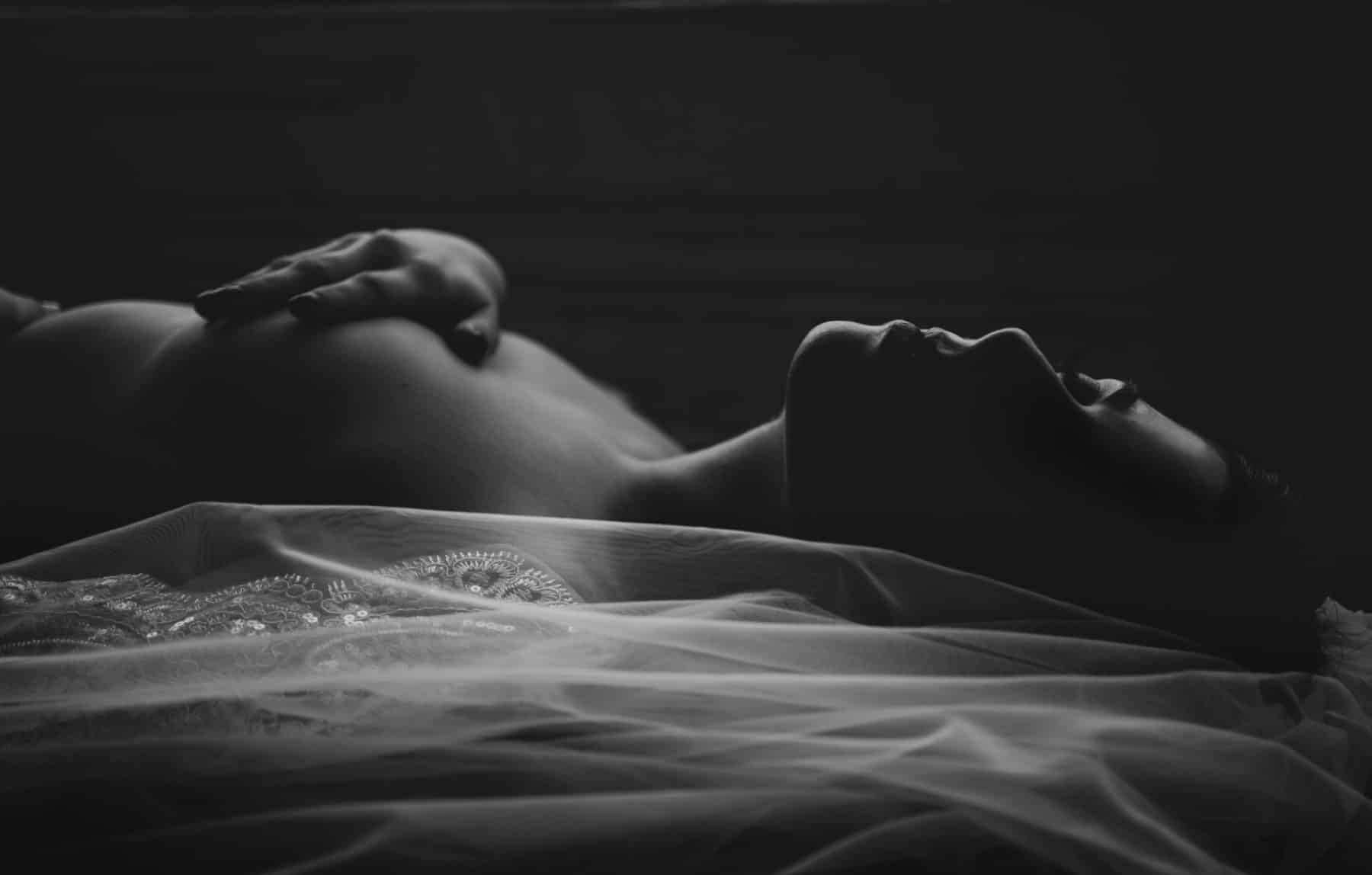 Black and white bridal boudoir portrait with veil, rim lighting from window, her on her back, eyes closed