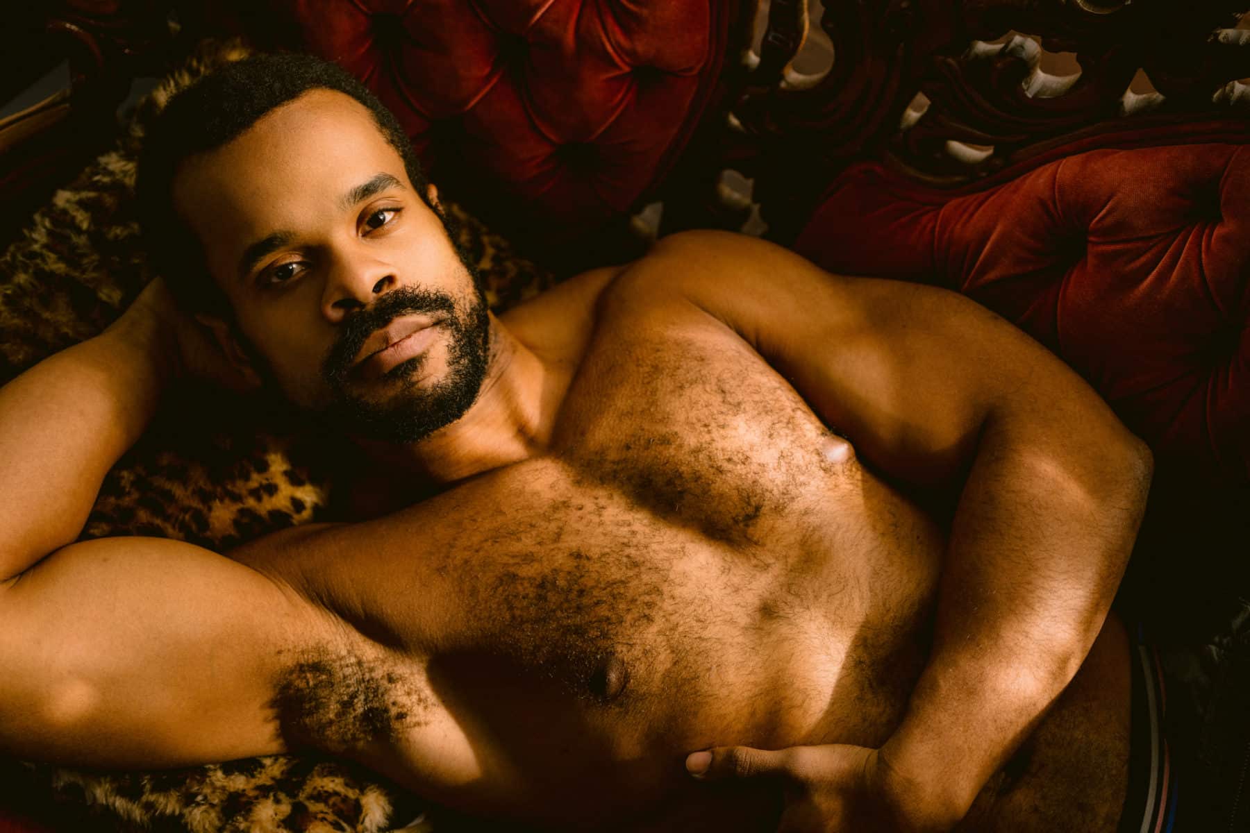 Men's boudoir photo of muscular black man, shirtless, laying on a red velvet couch, one arm behind his neck, looking right into the camera during his sexy dudoir session in philly suburbs